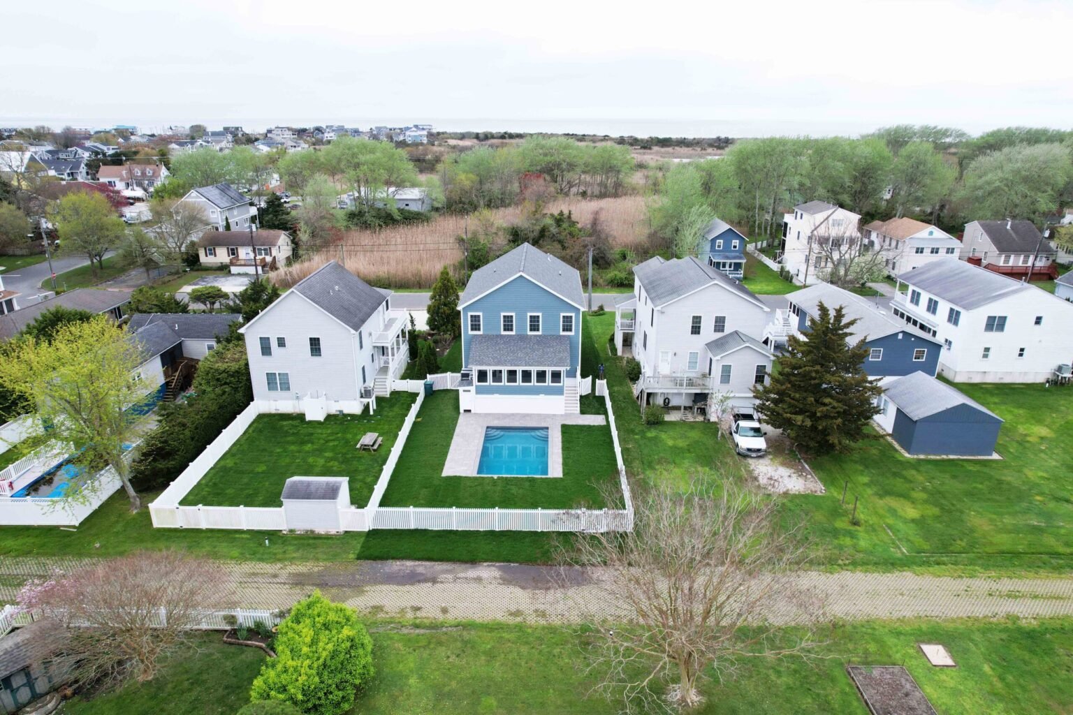 An aerial drone view of a rectangular in-ground pool with a paver surround set within a large grass backyard of a two-story blue-gray home. The yard is enclosed by a white vinyl fence, and the surrounding New Jersey neighborhood with mature trees and water