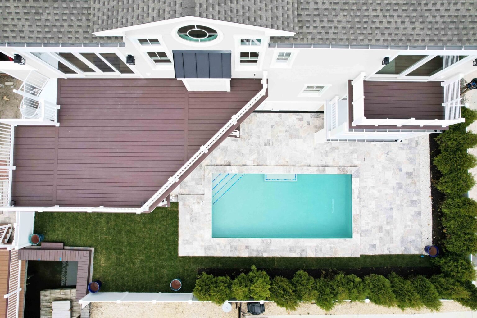Aerial view of a rectangular in-ground pool surrounded by a travertine paver patio, adjacent to a large dark-red composite wood deck with white railings. The property includes a manicured green lawn and is situated in a coastal residential neighborhood.