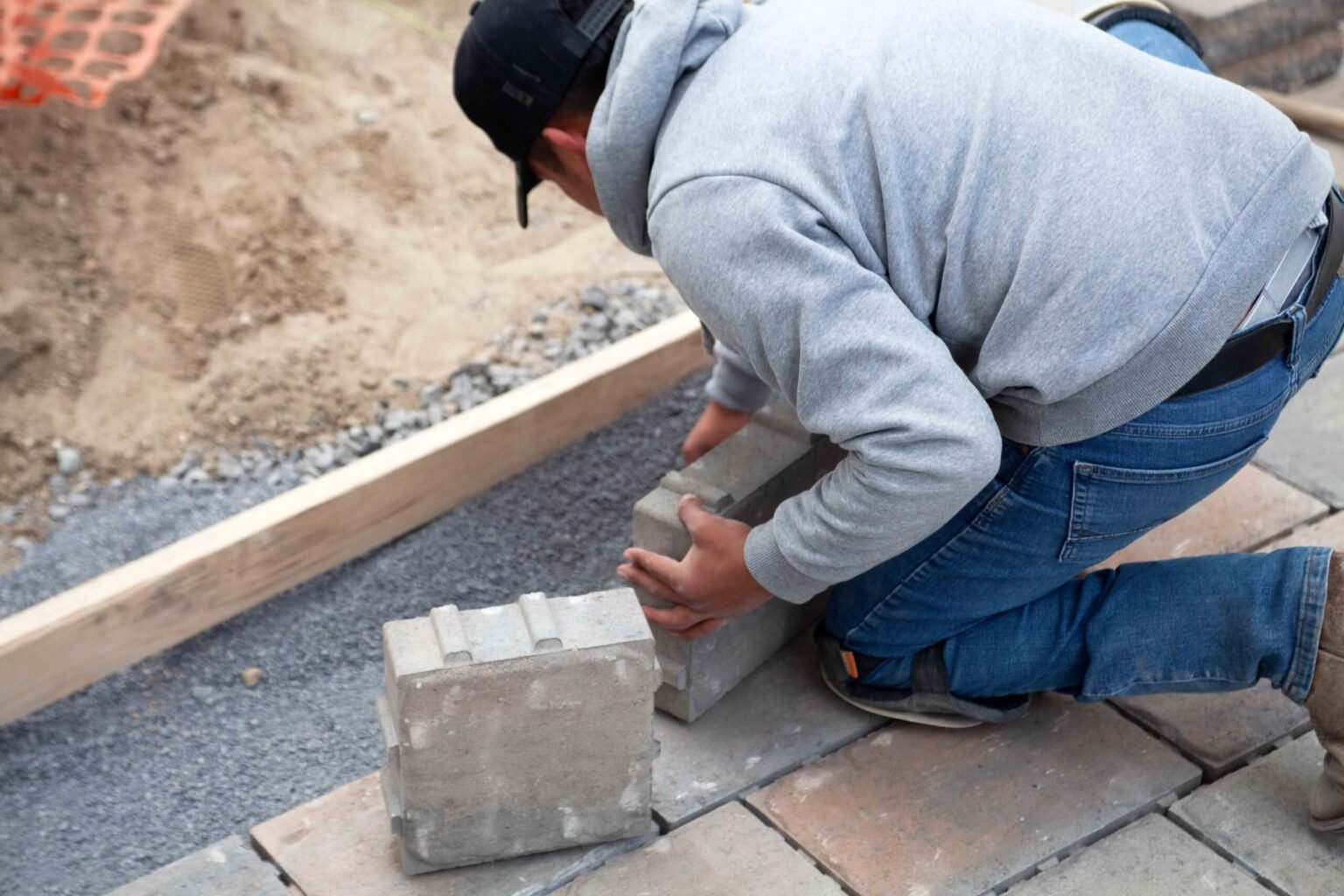 Construction phase photo showing a worker on hands and knees setting concrete paver blocks on a compacted gravel base during pool deck installation. A wood screed board, sand bed, and orange safety netting are visible in the background alongside partially 