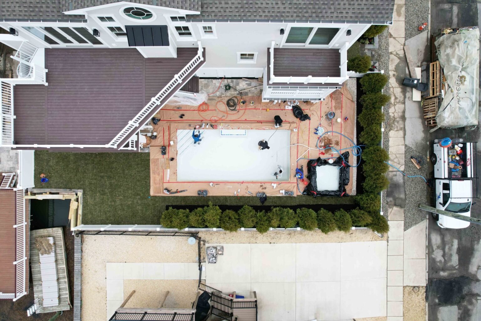Aerial construction photo shows a rectangular in-ground pool shell with a white gunite base being built in a compact backyard. Workers, hoses, and red string layout lines are visible on the surrounding paver deck area, with a multi-story house and dark woo