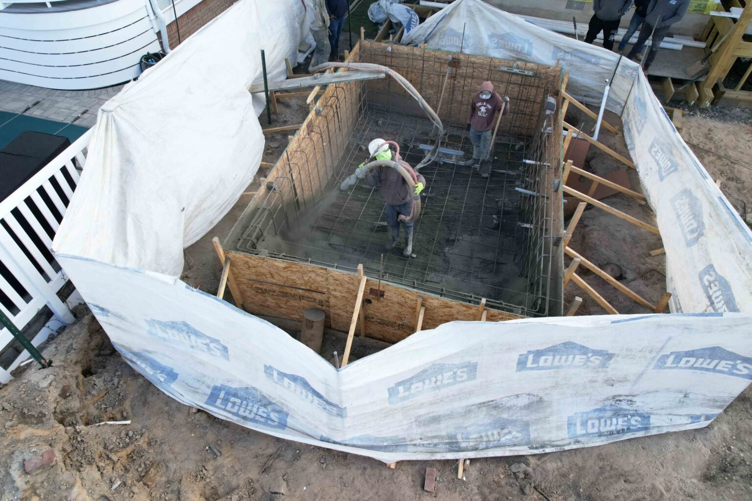 A construction phase photo showing two workers spraying gunite concrete into a rectangular pool shell reinforced with a steel rebar grid. The excavation site is surrounded by wood formwork and tarps. This is an in-progress pool construction photo.