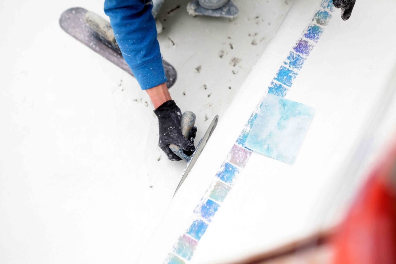 Close-up construction photo showing a worker in gloves using a trowel to set iridescent blue mosaic tiles along the waterline of an unfinished pool shell. The white plaster pool interior is visible with no water present.
