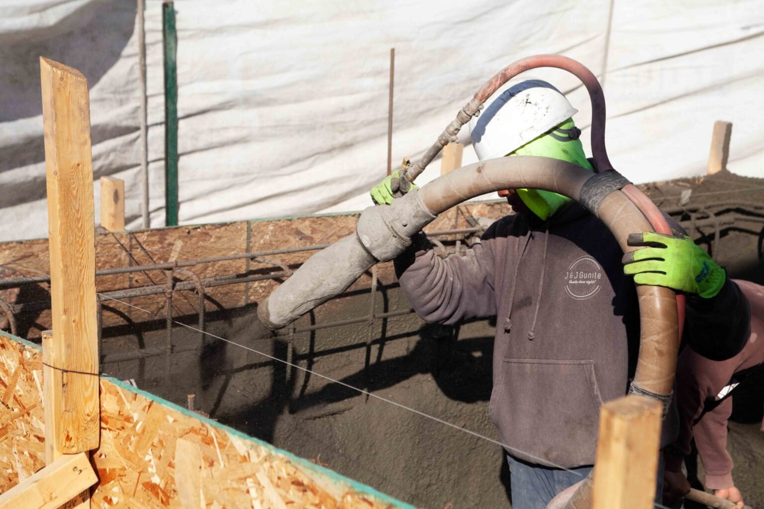 In-progress construction photo showing a worker in a hard hat and safety gear operating a shotcrete hose to spray gunite concrete onto the rebar shell of a pool under construction. Wooden form boards line the excavation perimeter, and a protective tarp cov