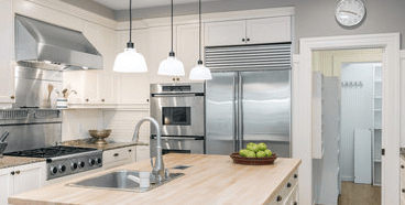 Indoor kitchen featuring white cabinetry, a butcher block island with a stainless steel sink, double wall ovens, a large stainless steel refrigerator, a gas range with stainless hood, granite countertops, and three pendant lights hanging from the ceiling.
