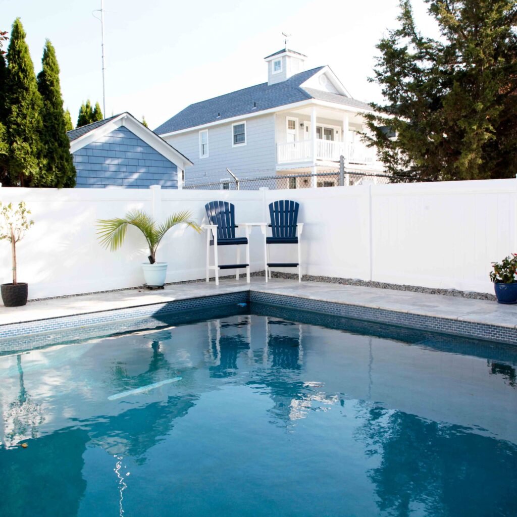 Rectangular in-ground pool with dark blue water and a blue mosaic tile waterline border, viewed from the water's edge. A gray stone or concrete patio deck runs along the far side, where two navy blue Adirondack bar chairs and a small white table are positi