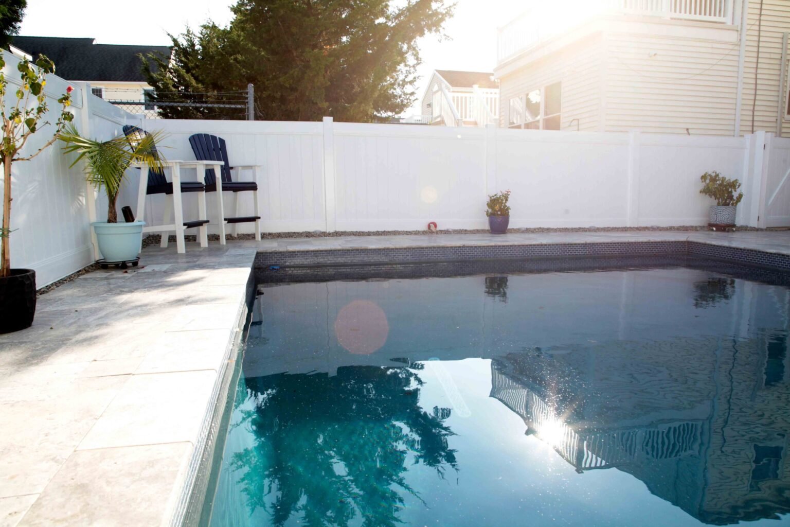 Rectangular in-ground pool with dark charcoal tile along the waterline, set in a compact backyard with a light stone or travertine paver deck. Two navy Adirondack bar-height chairs sit along the far deck edge. A white vinyl privacy fence encloses the yard,