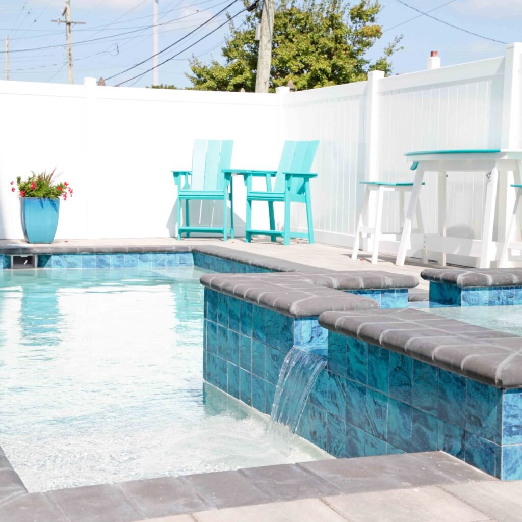 Close-up of an in-ground pool featuring a raised spa and sun shelf clad in blue iridescent square tile with gray cushioned seat pads and a sheet-style waterfall spillover into the pool. The surrounding deck is gray concrete pavers, with teal Adirondack cha