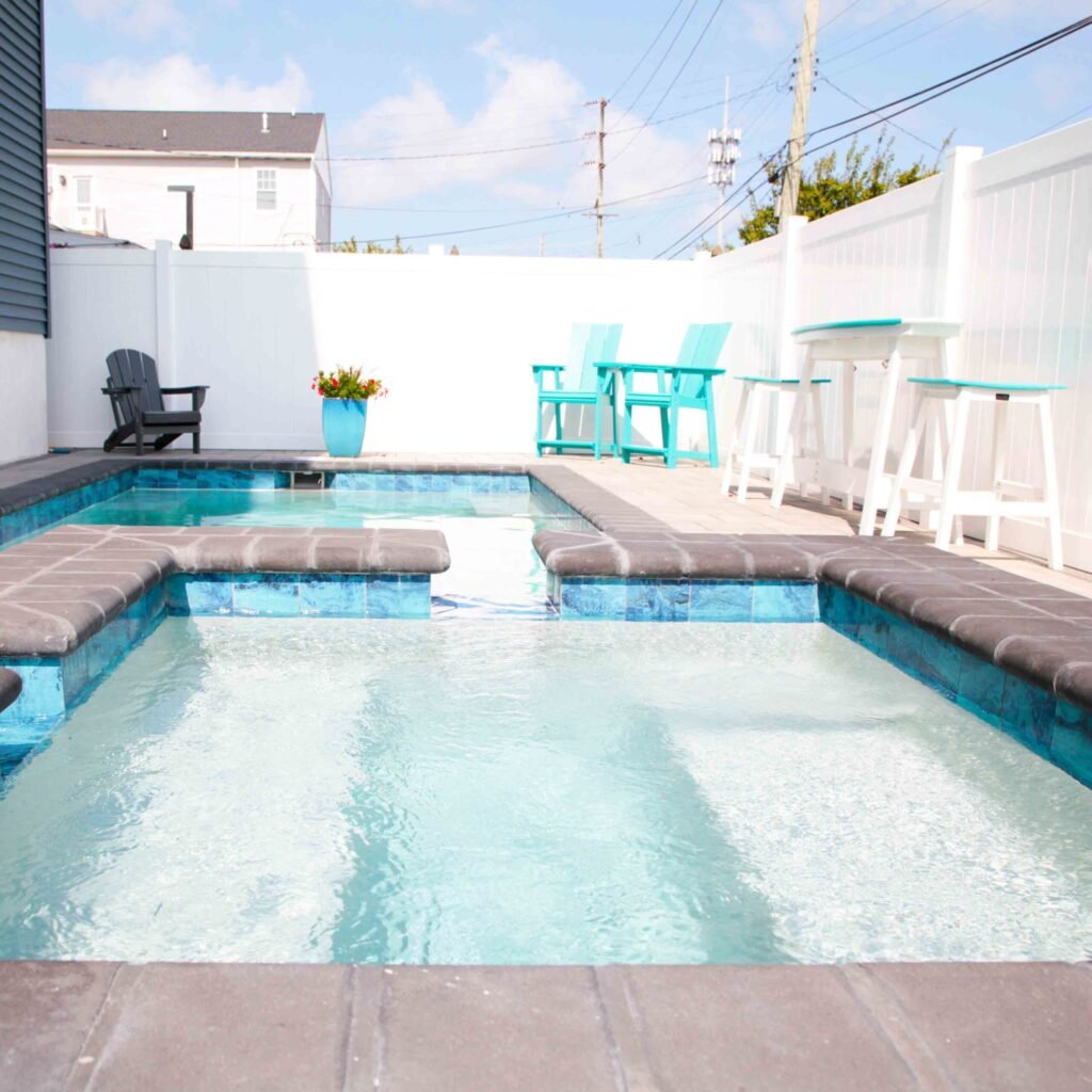 Rectangular in-ground pool viewed from deck level, with a raised spa and bench ledge clad in blue iridescent tile and capped with gray coping. The gray paver deck is furnished with teal and black Adirondack chairs and white bar stools, enclosed by a white 
