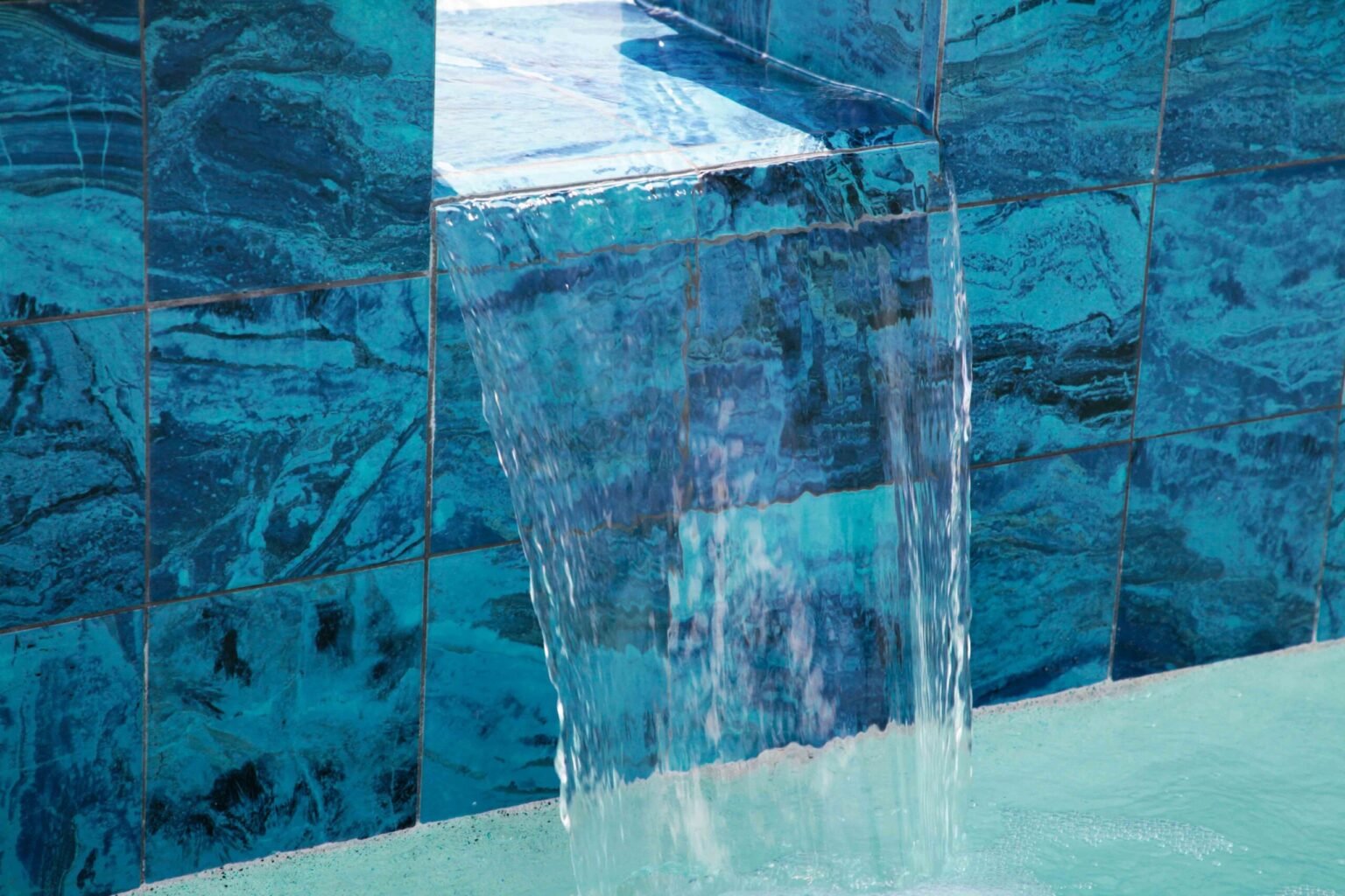 Close-up of a sheer descent waterfall spilling from a raised bond beam clad in large-format blue and teal marble-look porcelain tile into a pool below. Water streams in a thin sheet down the tile face, with the pool surface visible at the bottom of the fra