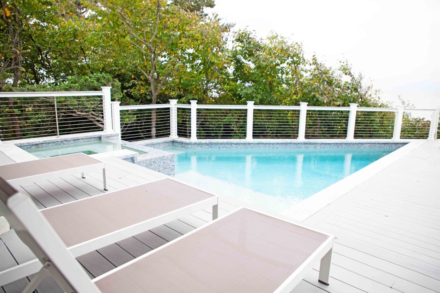 Rectangular in-ground pool with an attached spa in the far corner, set on a composite wood deck with white cable-rail fencing along the perimeter. Three lounge chairs are positioned in the foreground, and mature trees are visible beyond the fence line.