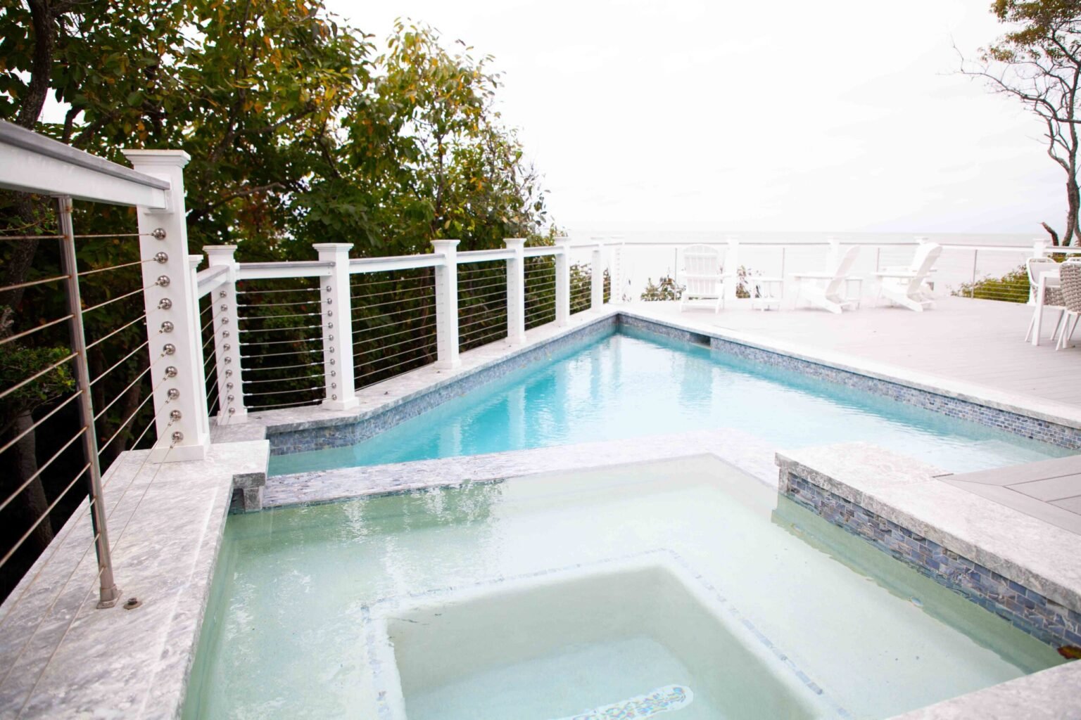L-shaped in-ground pool with an attached square spa, both finished in blue-gray mosaic tile with white marble coping, on a white marble deck enclosed by stainless steel cable railings; open bay water and white lounge chairs are visible in the background.