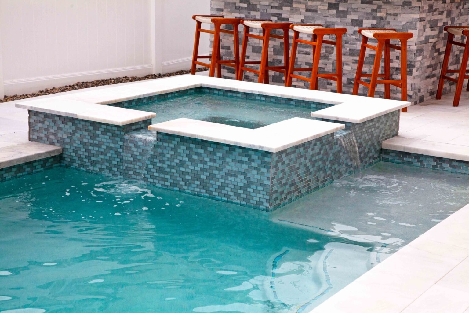 Square in-ground spa tiled in blue-gray glass mosaic with white marble coping overflows into an adjacent rectangular pool; wooden bar stools line a stacked-stone accent wall in the background on a white paver deck.
