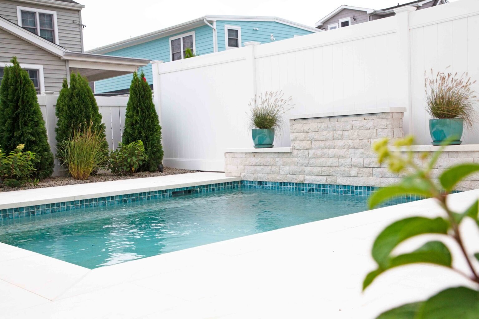 A small rectangular in-ground pool with a teal mosaic tile band along the waterline and white limestone coping. A stacked-stone spillway wall with a broad flat cap anchors the rear corner, topped with two teal ceramic planters holding ornamental grasses. A