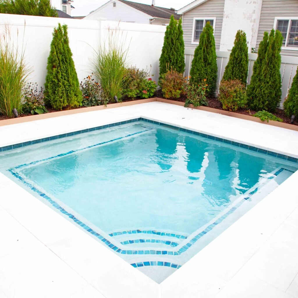 Small rectangular in-ground pool with light blue water, blue mosaic tile border, and fan-shaped entry steps along the near edge. The white concrete surround is bordered by a raised planting bed with columnar arborvitae, ornamental grasses, and flowering sh