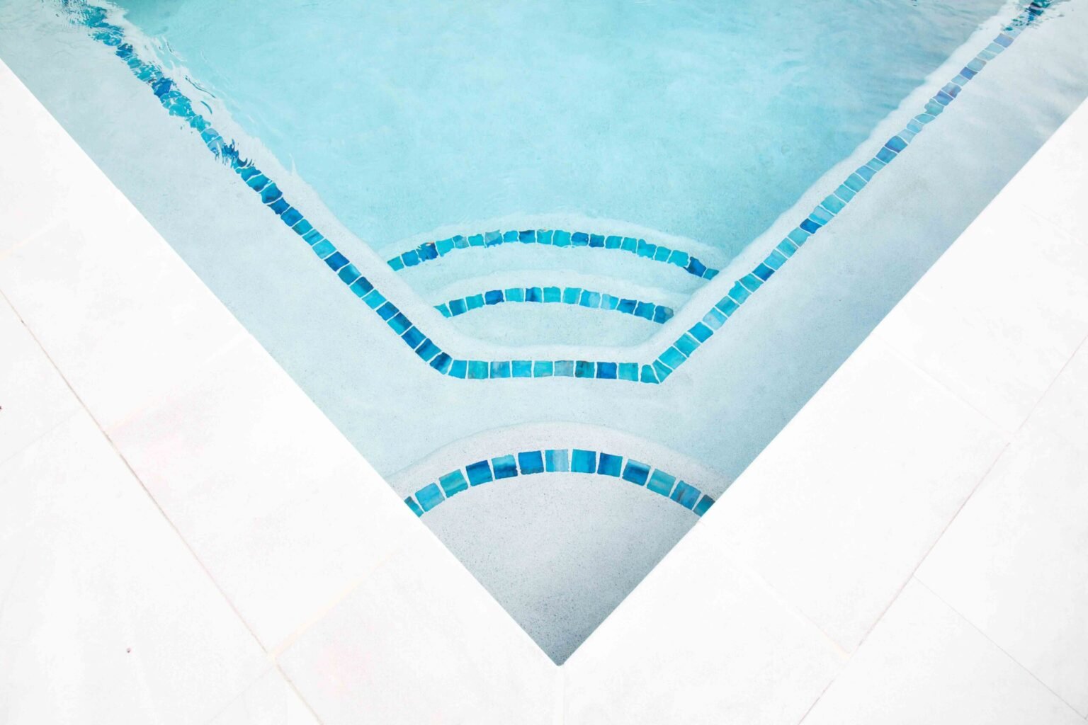 Close-up of an in-ground pool corner showing fan-shaped submerged entry steps with blue mosaic tile accent bands lining each step tread. The pool interior is light gray plaster, and white coping borders the pool edge.