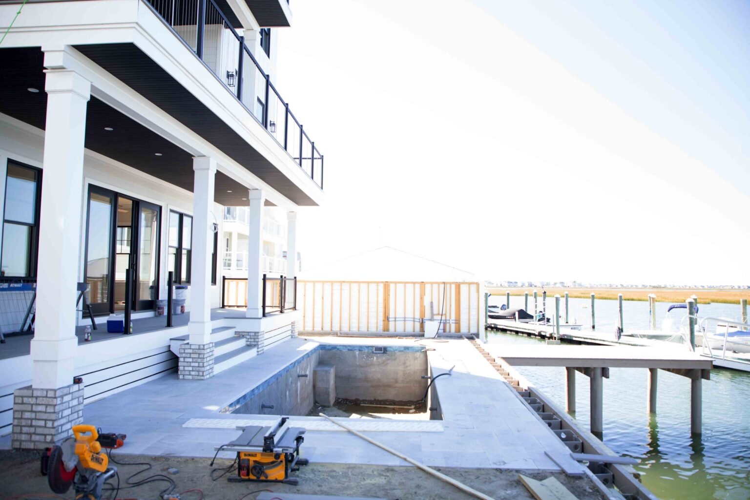 In-ground pool shell under active construction on a waterfront property. The gunite shell is exposed and unfilled, surrounded by partially installed gray stone pavers. DeWalt power tools and saw equipment are visible on the patio. A boat dock with pilings 