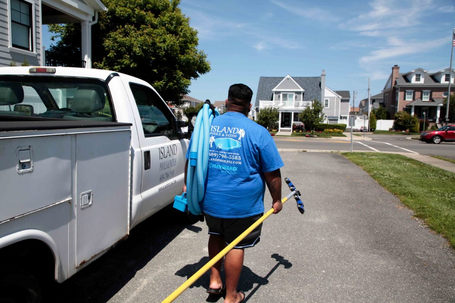 A pool service technician wearing an Island Pools and Patios branded blue t-shirt carries a yellow telescoping pole and a blue vacuum hose toward a white company service truck parked on a residential street. The truck door displays the Island Pools and Pat