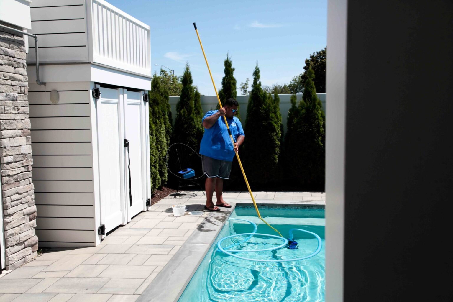 A pool service technician in a blue shirt uses a long-handled vacuum pole to clean a rectangular in-ground pool from a large-format paver patio deck, with a white lap-siding pool house with stone veneer accent and a row of arborvitae in the background.