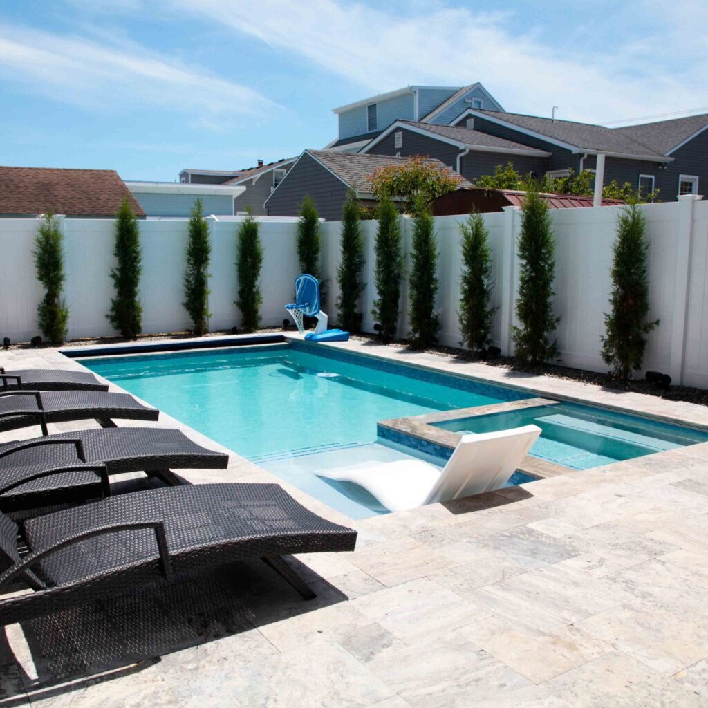 Rectangular in-ground pool with blue water and an attached raised spa, set on a travertine paver deck. A white tanning ledge with a lounger is visible at the shallow end. Dark wicker chaise lounges are arranged poolside, and a row of columnar evergreen tre