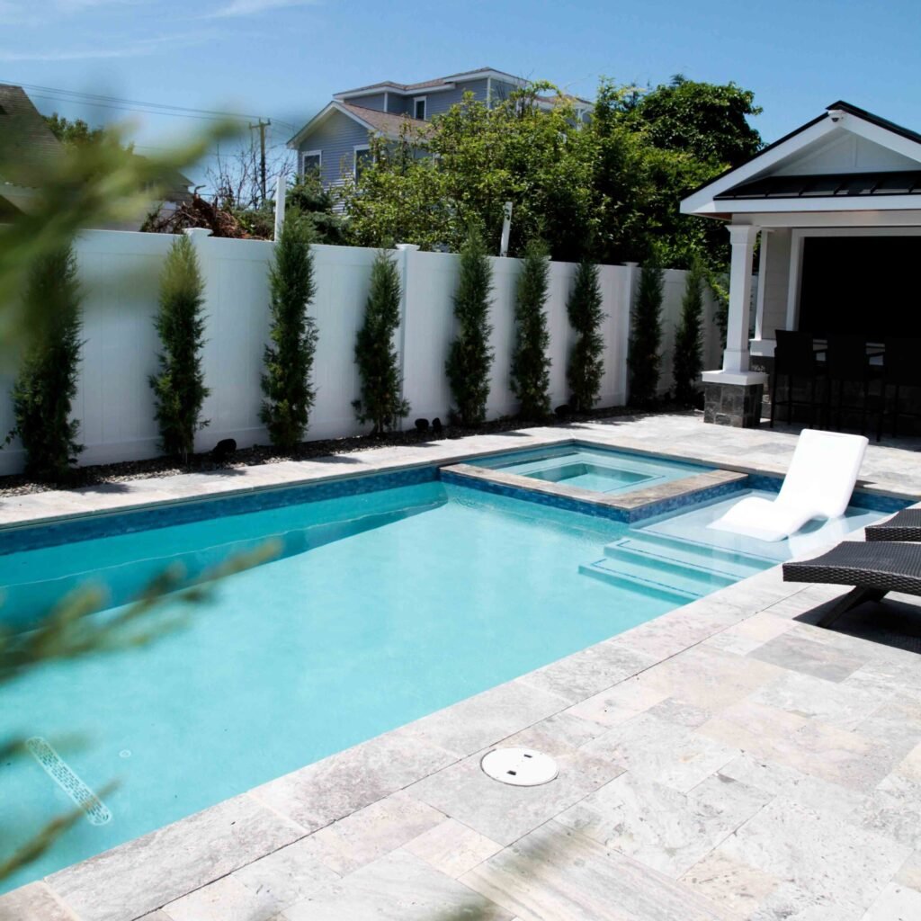 Rectangular in-ground pool with clear blue water and an adjacent elevated square spa, set on a travertine paver surround with light gray coping. A white vinyl privacy fence lined with tall columnar evergreens borders the yard, and a white pergola structure