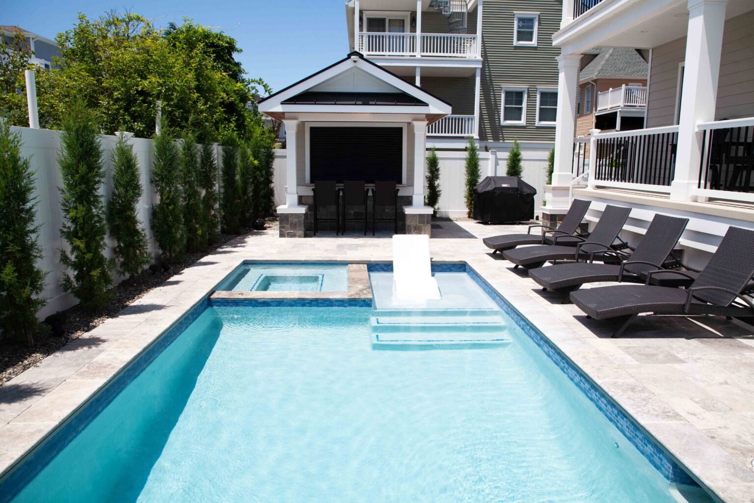 A rectangular in-ground pool with a raised attached spa sits on a travertine paver deck lined with dark wicker lounge chairs; a white-painted pergola cabana with an outdoor television and bar seating anchors the far end, enclosed by a white vinyl privacy f