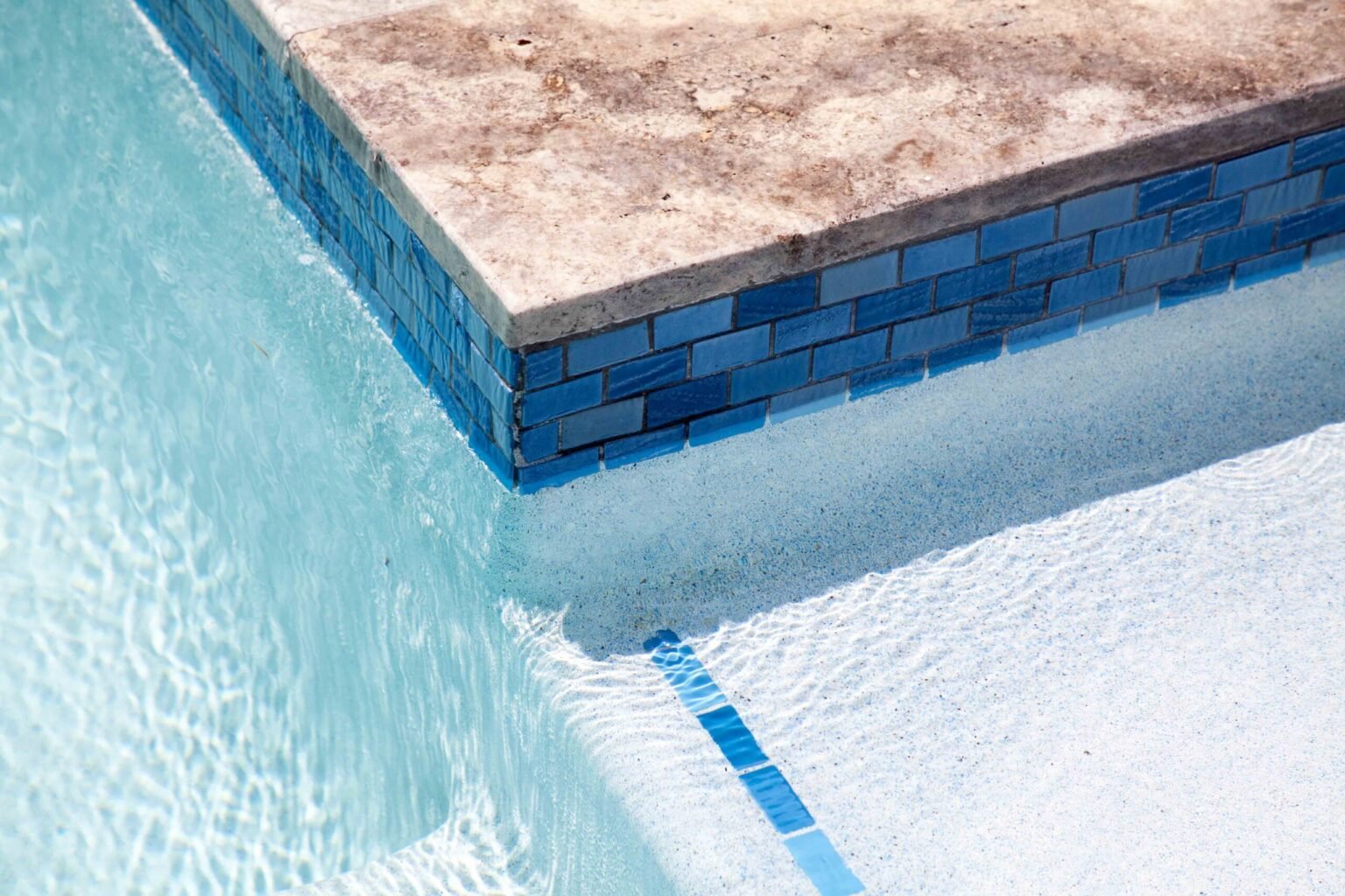 Close-up corner view of an in-ground pool showing a course of rectangular blue glazed tile along the waterline beneath a travertine stone coping cap, with white plaster visible on the pool floor below the water surface.