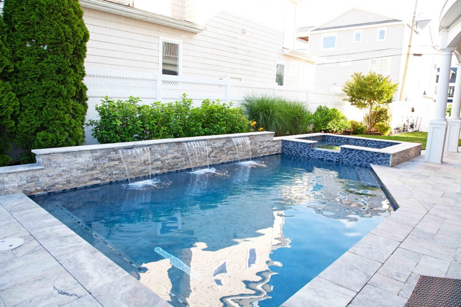 Geometric in-ground pool with a stacked-stone sheer descent waterfall along one wall, with water cascading across the full width into the pool. An elevated square spa with dark blue tile sits at the far end, and the surrounding deck is finished in large-fo