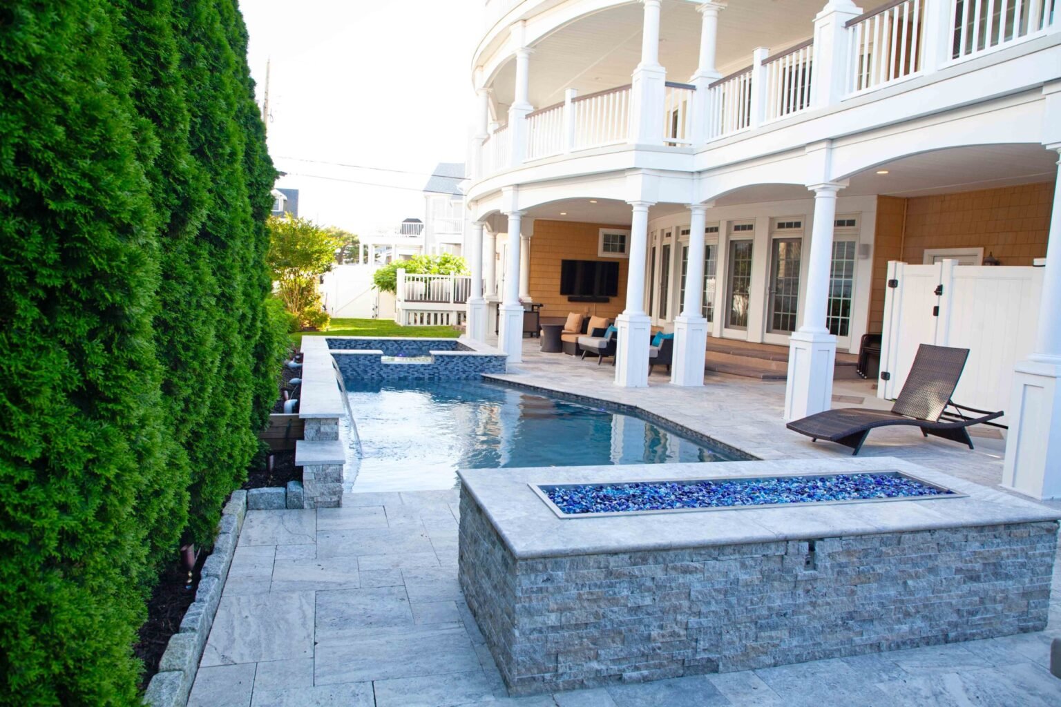 A narrow rectangular lap pool with blue glass tile trim runs along a travertine paver patio beside a two-story home with a curved wraparound covered porch supported by white columns. A rectangular raised fire pit filled with blue glass is in the foreground
