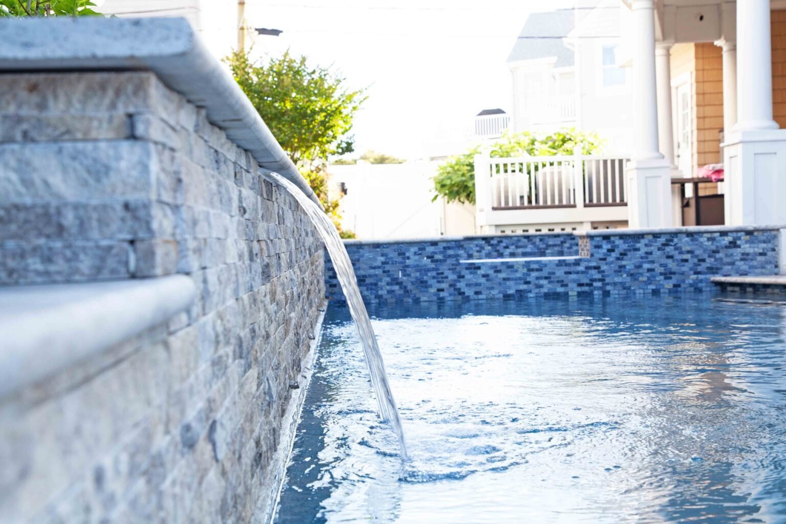 Close-up of a raised stone veneer retaining wall with a sheet waterfall cascading into a pool. The pool interior is lined with dark blue glass mosaic tile visible along the waterline. A white porch with columns and a vinyl fence are visible in the soft-foc