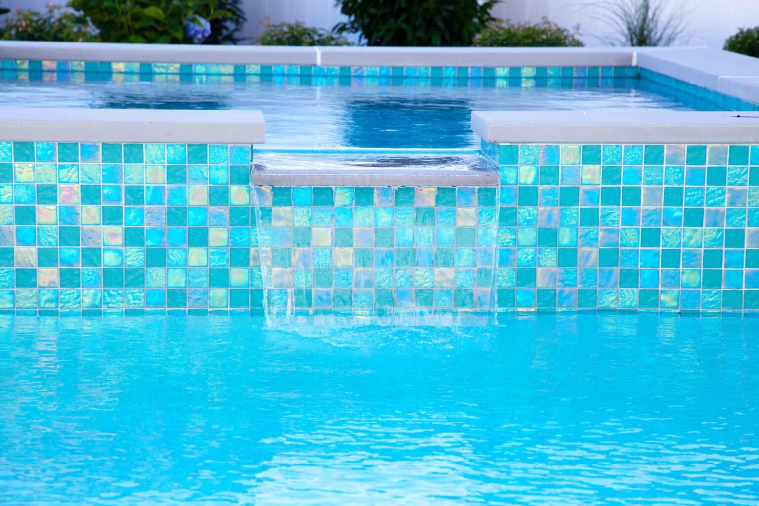 Close-up of a raised spa with an iridescent teal and aqua glass mosaic tile face, water spilling over a ledge spillway into the rectangular pool below, with a white vinyl fence and flowering plants visible in the background.