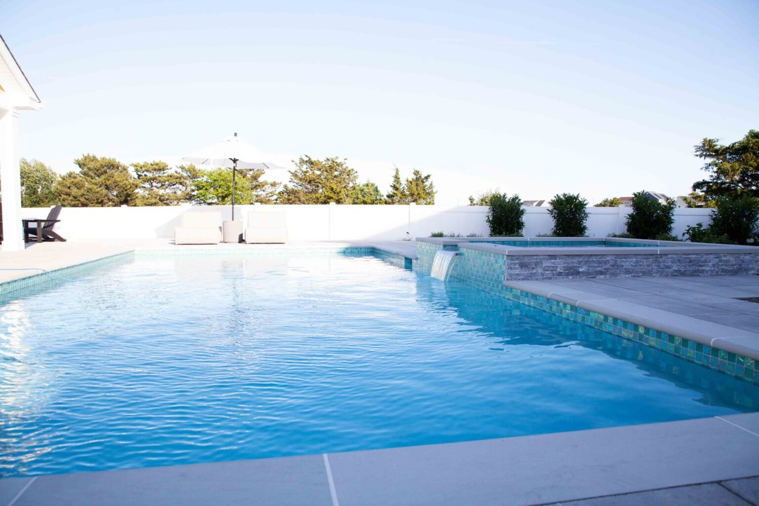 A completed rectangular in-ground pool with a light gray concrete coping surround and iridescent green mosaic waterline tile. An elevated spa with stacked stone veneer and a sheet-water spillover feature is positioned at the far end. The pool deck is finis