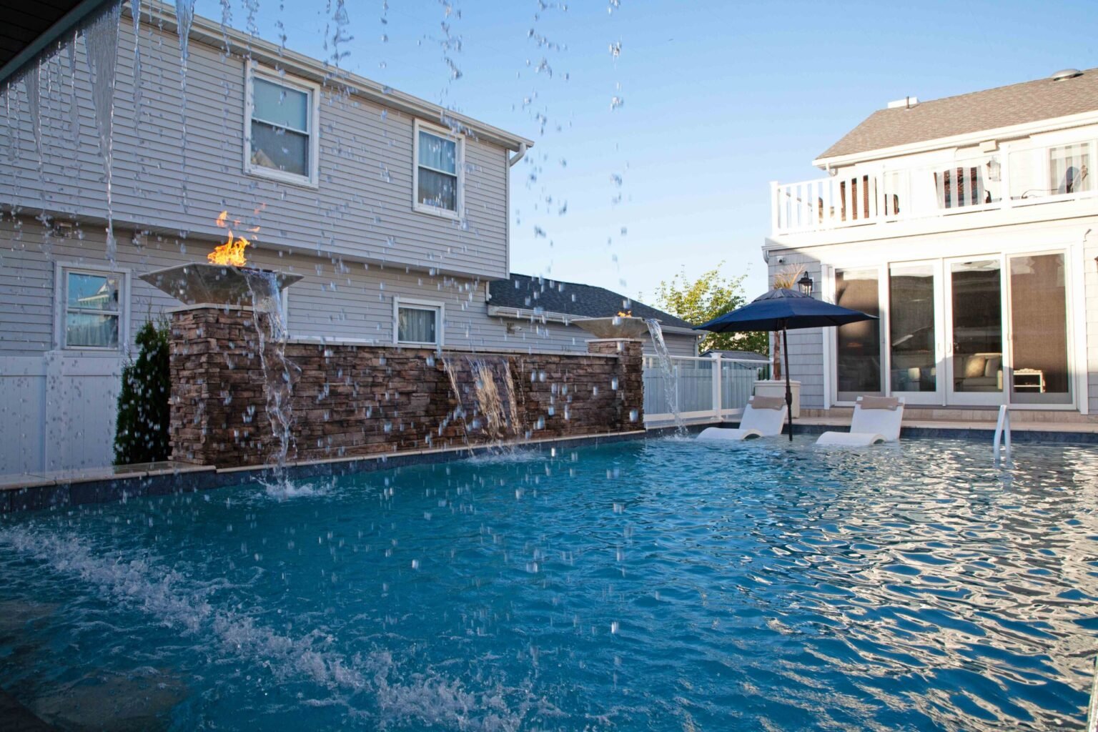 A large in-ground pool features a raised stacked-stone water wall with two active fire bowls and multiple sheer descent water curtains cascading into the pool; white sun loungers and a navy umbrella are positioned on the surrounding concrete deck beside a 