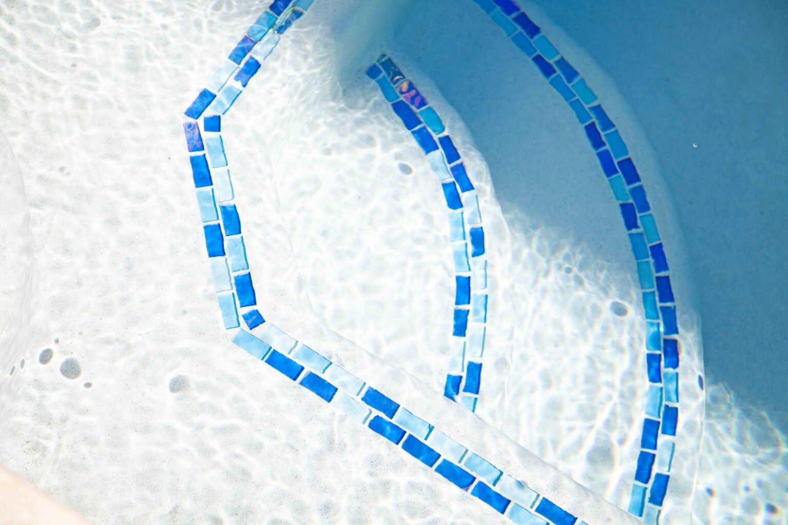 Close-up underwater view of curved pool entry steps outlined with blue and light-blue glass mosaic tile strips set against a white plaster pool interior. The water is clear and the tile reflects light in an iridescent pattern.
