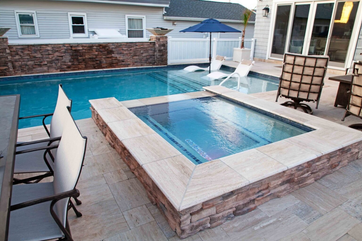 Rectangular in-ground pool with blue water and a dark tile waterline band, adjacent to an elevated square raised-bond-beam spa clad in stacked stone veneer with a travertine coping cap. The travertine paver deck is furnished with sling chairs and a patio u