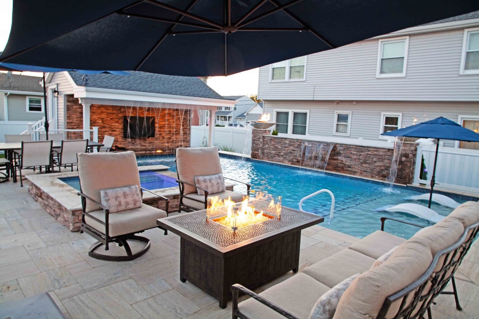 Rectangular in-ground pool with a raised stone waterfall wall along the far edge, flanked by torch-style sconces. A travertine paver patio surrounds the pool with patio seating and an outdoor propane fire pit table in the foreground. A pool house with ston