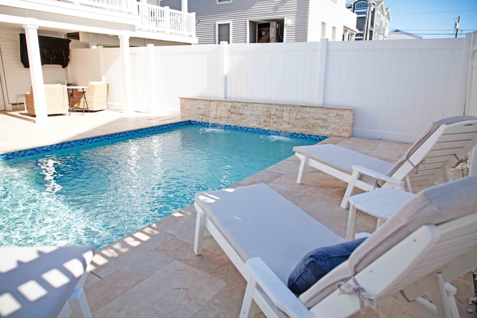 Rectangular in-ground pool with blue water and a natural stone veneer spillover wall at the far end, surrounded by a travertine paver deck with white vinyl privacy fence. Two white lounge chairs sit poolside, and a covered patio with outdoor seating is vis