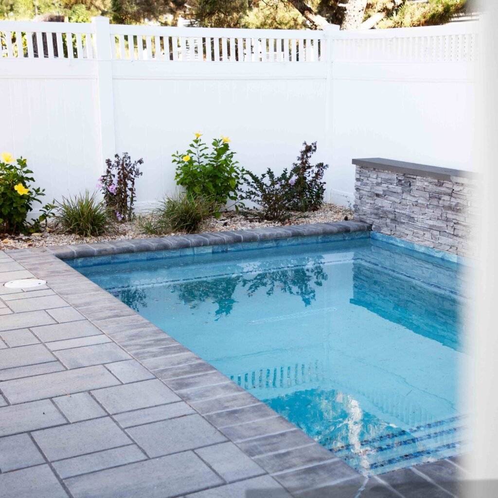 A small rectangular in-ground pool surrounded by a gray paver deck. A stacked-stone accent wall with a dark-capped ledge serves as a waterfall feature at the far end. A white vinyl privacy fence encloses the yard, with a garden bed of flowering plants and 