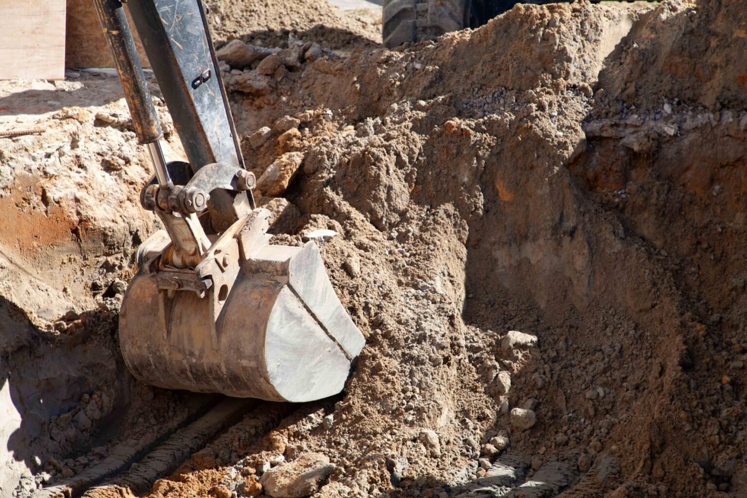 A construction photo shows an excavator bucket actively digging into rocky clay soil during the pool excavation phase. The walls of the dig reveal layered earth and rock, with no pool shell or water present. This is an early-stage pool construction process