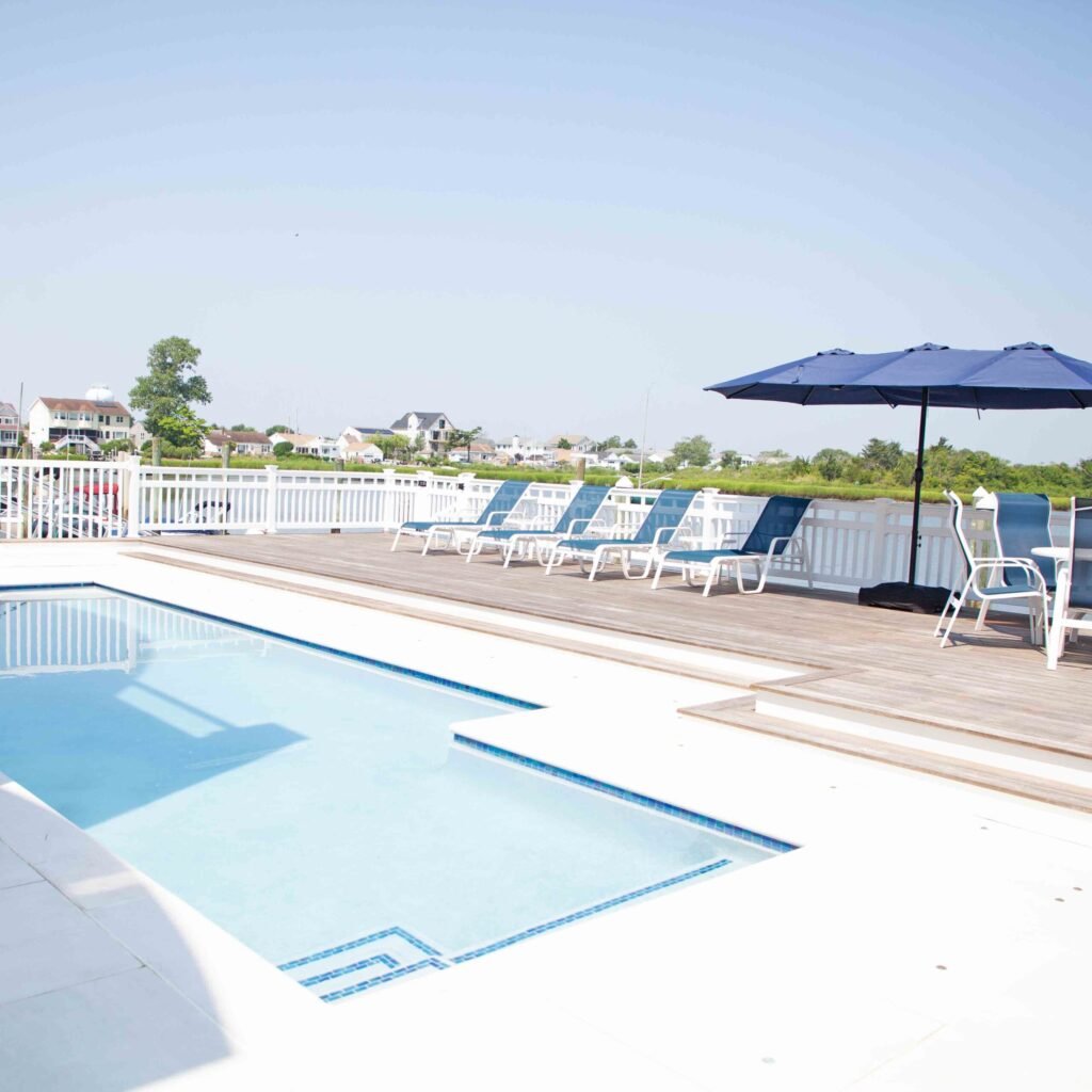 Rectangular in-ground pool with light blue water and blue mosaic tile inlay sits on a wood plank deck surrounded by white railing fence, with lounge chairs, a navy market umbrella, and an open bay waterway visible in the background.
