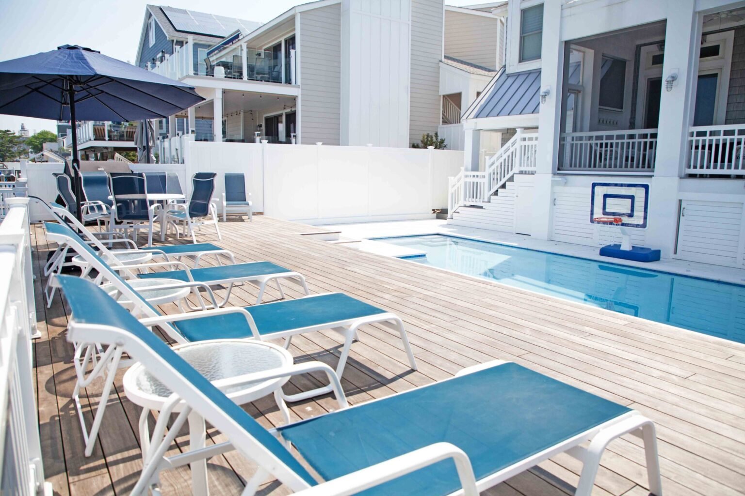 Rectangular in-ground pool on an elevated wood deck surrounded by blue lounge chairs and a patio umbrella with dining seating. A pool basketball hoop is mounted in the water, and a white vinyl fence encloses the space with a coastal neighborhood in the bac