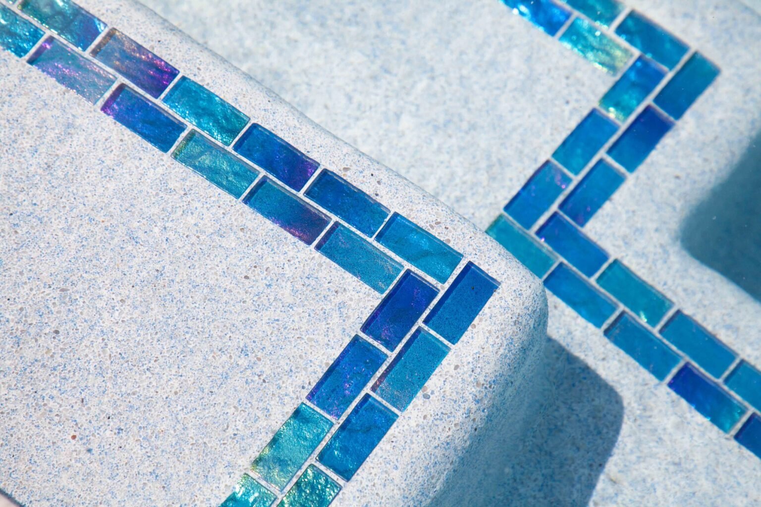 A close-up detail of a pool interior step and waterline, showing a row of iridescent blue and teal glass mosaic tiles set against a light gray concrete coping. The tiles shift between blue, purple, and green tones, and a second tile band is visible on an a