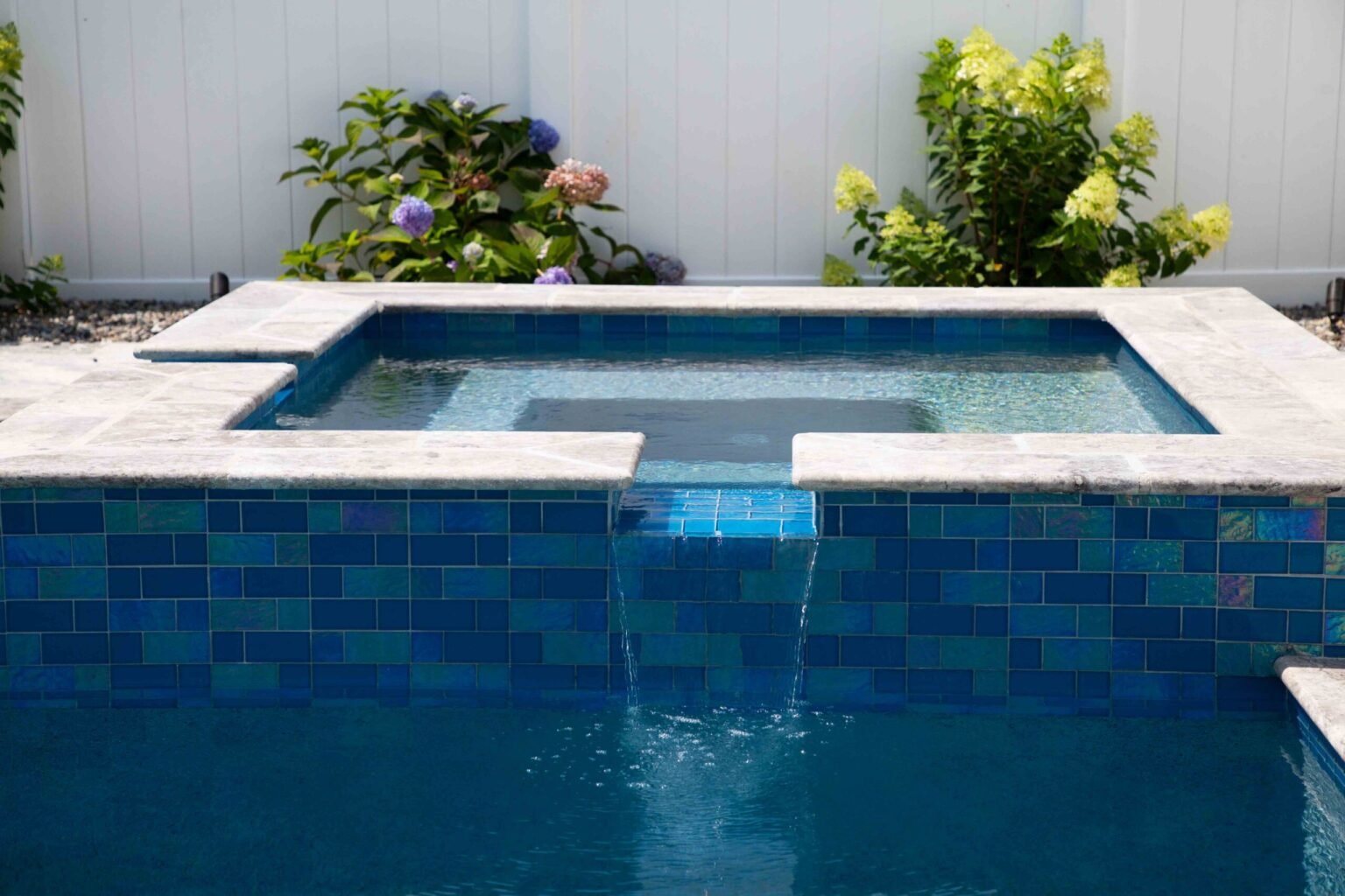 A completed square raised spa with a gray travertine coping surround and exterior walls fully clad in large-format iridescent blue and teal mosaic tiles. Water spills over a central weir into the pool below. Hydrangea shrubs in bloom are planted along a wh
