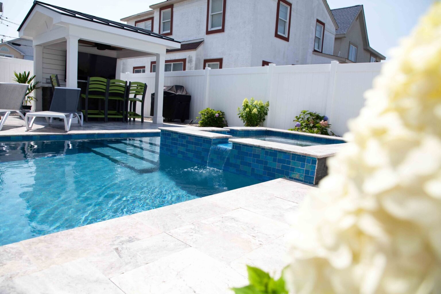 A rectangular in-ground pool with an attached raised spa and blue tile spillover waterfall is set on a travertine paver deck; a covered pergola with ceiling fan, dining chairs, and a gas grill occupies the background, enclosed by a white vinyl privacy fenc