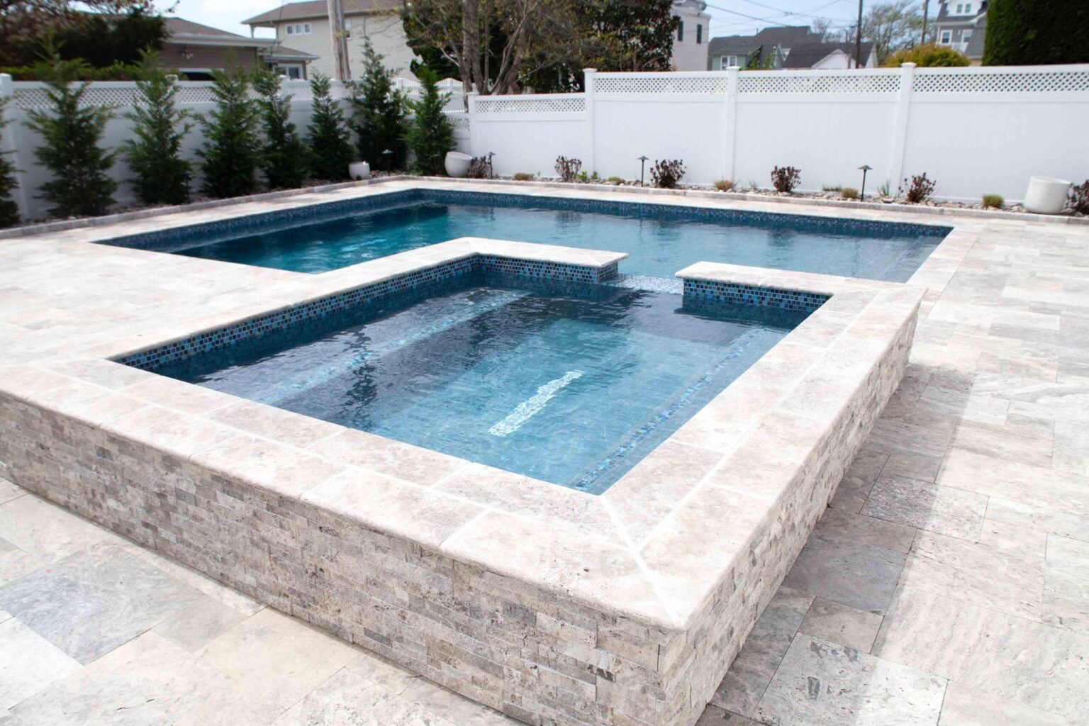 A geometric in-ground pool with an attached raised square spa, both finished with iridescent blue glass tile trim. The surround and raised spa walls are clad in stacked natural travertine stone, and the pool deck is laid in large-format travertine pavers. 