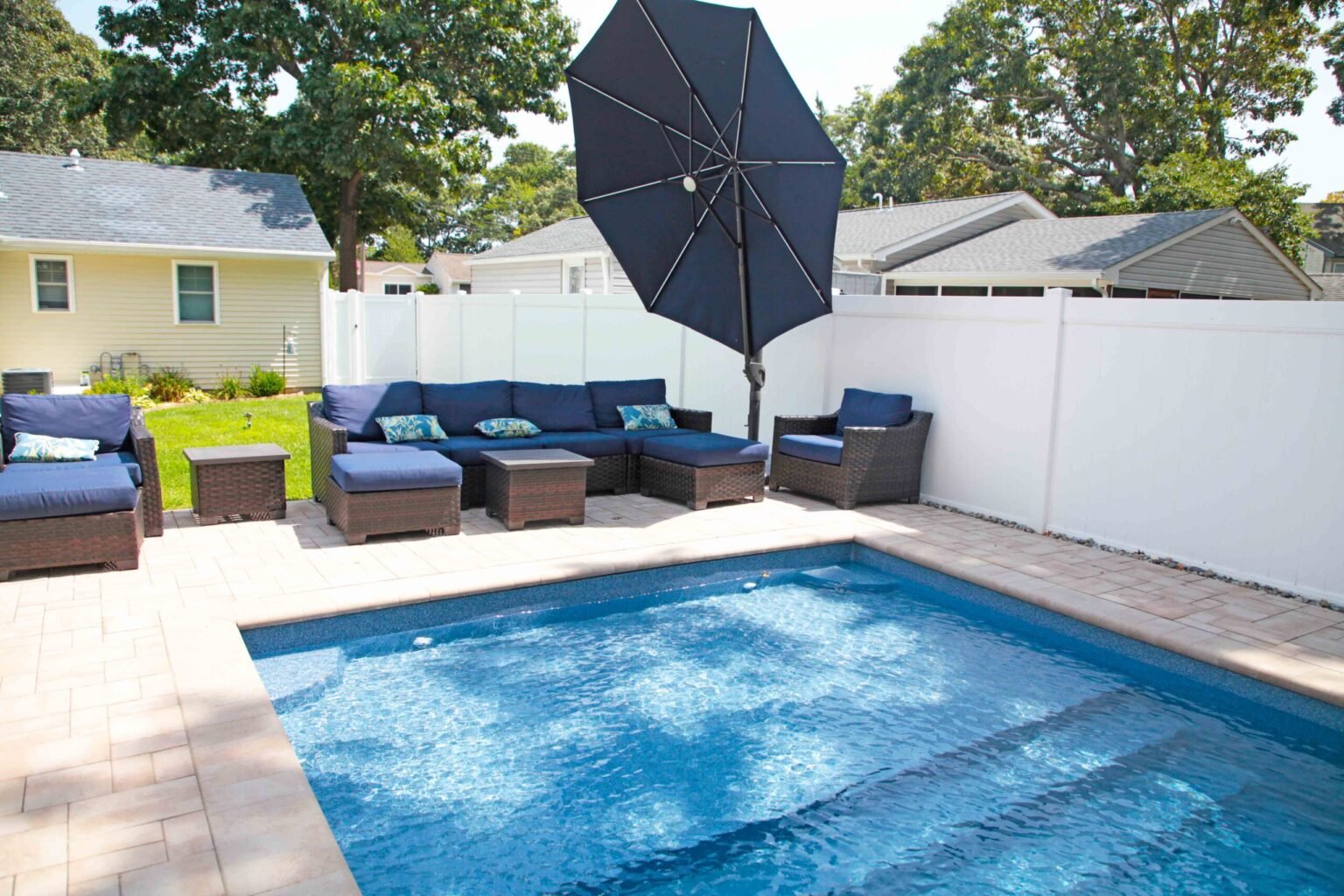 A small rectangular in-ground pool with tan paver coping and deck surround is shown from pool level. A navy blue wicker sectional sofa set and large black cantilever umbrella occupy the patio. A white vinyl privacy fence encloses the suburban backyard, wit