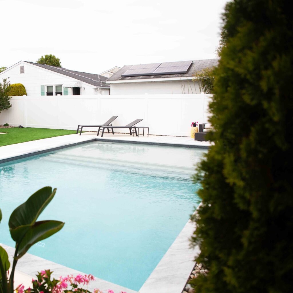 Rectangular in-ground pool with light blue water and dark gray waterline tile, framed by white concrete coping. Two dark chaise lounges sit on the pool deck near a white vinyl privacy fence. Green lawn, tropical foliage in the foreground, and a white house