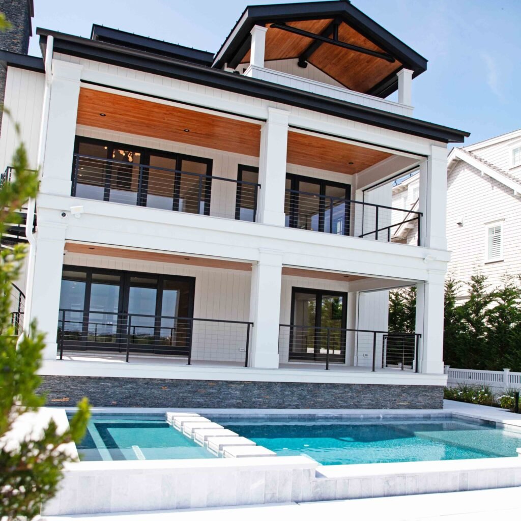 Rectangular in-ground pool with a raised attached spa section featuring white stone coping and a dark stacked-stone surround, set against a two-story coastal home with covered porches, wood-plank ceilings, and black cable railings.