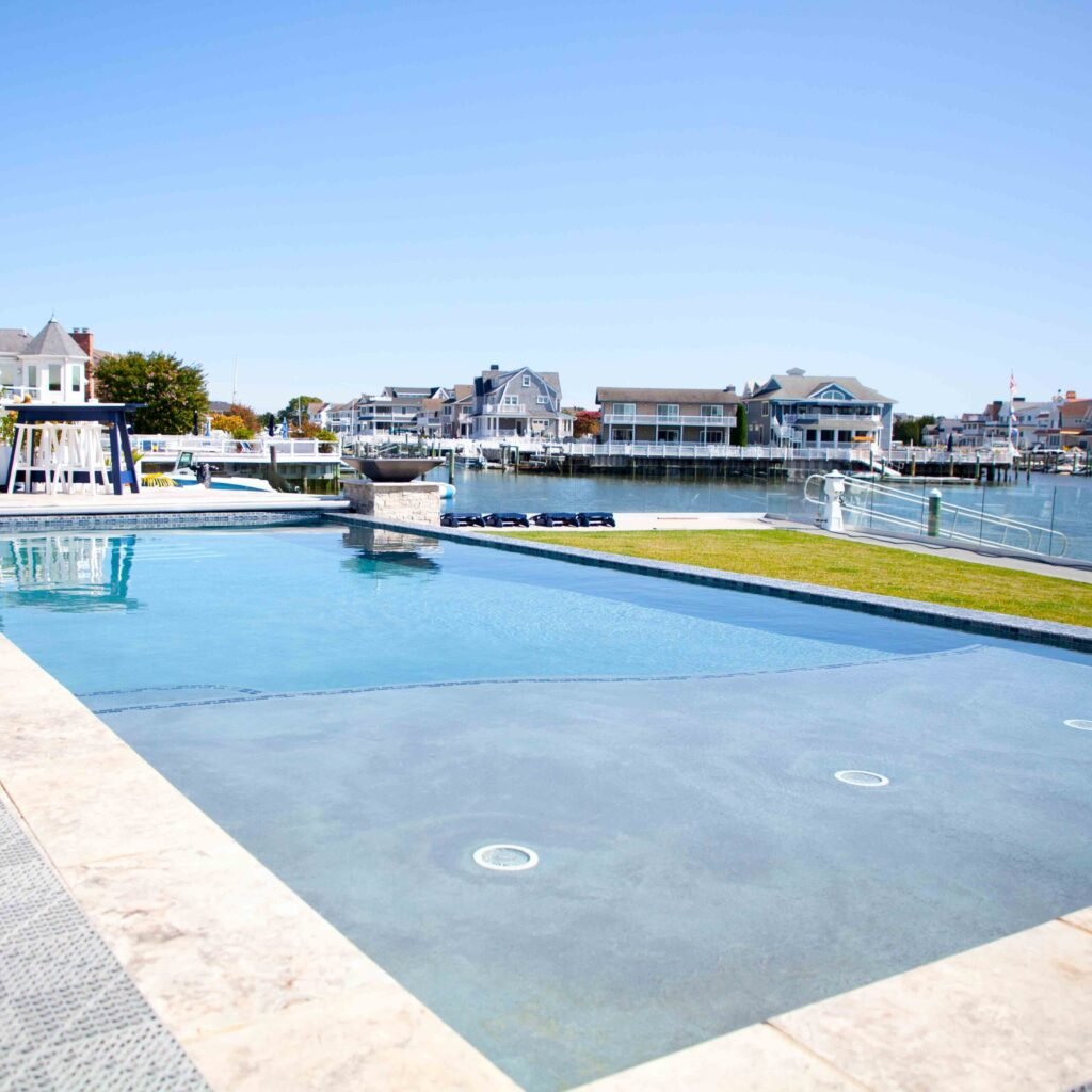 A large rectangular in-ground pool with a vanishing-edge design overlooking a marina and bay. The pool features gray plaster finish, recessed underwater lighting, and travertine coping. A raised stone pedestal with a fire bowl sits at the far end, and a bo